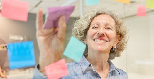 Eine Frau steht an einer Glaswand und heftet ein Post-it dran. Dabei lächelt sie in die Kamera